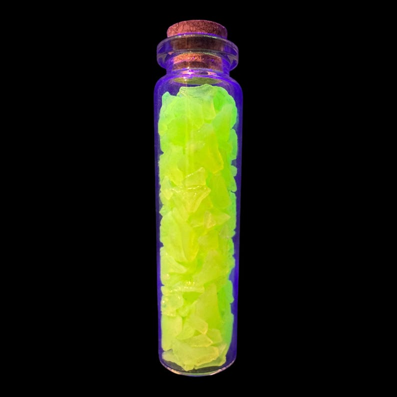 K&ouml;nnte beinhalten: Eine klare Glasflasche mit Korken, gef&uuml;llt mit hellgr&uuml;nen, unregelm&auml;&szlig;ig geformten St&uuml;cken. Die Flasche hat einen violetten Farbton an den R&auml;ndern, vor einem schwarzen Hintergrund.