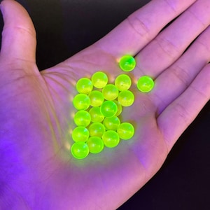 May include: A collection of small, spherical, translucent beads that glow with a vibrant neon yellow-green colour. These round objects are smooth and uniform in shape, appearing to emit a bright light against the dark background of the palm.
