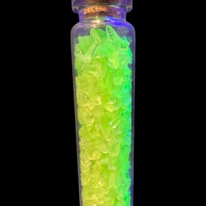 K&ouml;nnte beinhalten: Ein kleines, klares Glasfl&auml;schchen gef&uuml;llt mit leuchtend gr&uuml;nen und gelb-gr&uuml;nen Fragmenten. Die Flasche hat einen Korken und steht vor einem schwarzen Hintergrund. Der Inhalt scheint zu leuchten.