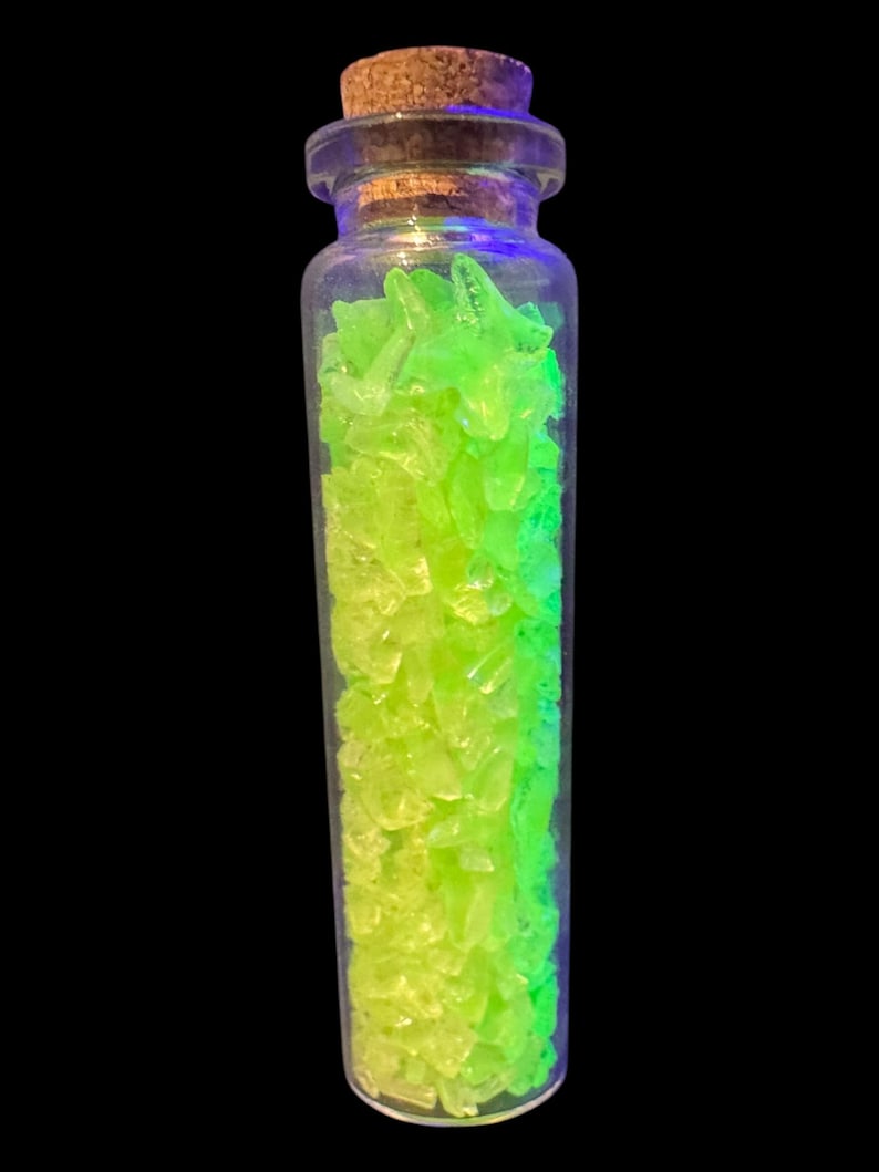 K&ouml;nnte beinhalten: Ein kleines, klares Glasfl&auml;schchen gef&uuml;llt mit leuchtend gr&uuml;nen und gelb-gr&uuml;nen Fragmenten. Die Flasche hat einen Korken und steht vor einem schwarzen Hintergrund. Der Inhalt scheint zu leuchten.