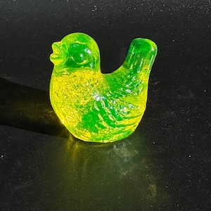 Puede incluir: Una figura de pájaro de cristal verde translúcido. El pájaro tiene un cuerpo redondeado y una cola curvada. El cristal tiene un aspecto texturizado, con tonos amarillos y verdes. El pájaro está sobre una superficie oscura.