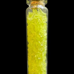 K&ouml;nnte beinhalten: Ein kleines, klares Glasfl&auml;schchen mit zahlreichen hellgelb-gr&uuml;nen, fragmentierten St&uuml;cken. Die Flasche ist mit einem Korken verschlossen. Der Hintergrund ist schwarz und hebt den Inhalt der Flasche hervor.