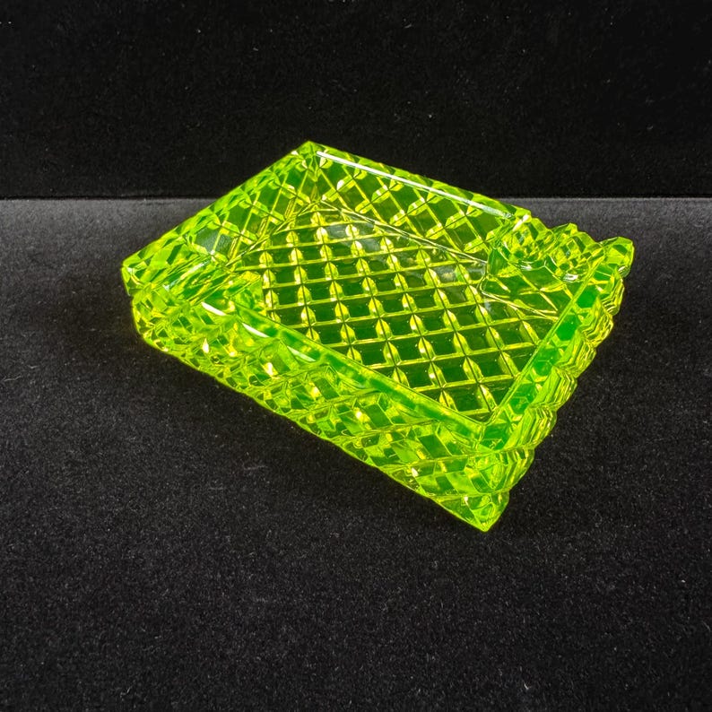 May include: A neon green ashtray with a diamond pattern. The ashtray is rectangular with a slightly raised edge and a small notch on one side. The color is a bright, vibrant green, and the ashtray appears to be made of glass.