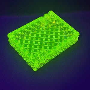 May include: A neon green, rectangular ashtray with a diamond pattern. The ashtray has a small, raised area for resting cigarettes. The glass ashtray is a vibrant, eye-catching piece.