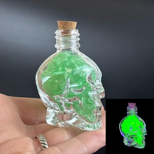 Peut inclure: Une bouteille en verre transparent en forme de crâne avec un bouchon en liège, remplie de petits éclats de cristal vert translucide. L'image encartée montre la même bouteille crâne brillant d'une lumière vert néon dans l'obscurité.