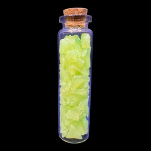 K&ouml;nnte beinhalten: Eine kleine, klare Glasflasche gef&uuml;llt mit hellen, neon-gr&uuml;nen, unregelm&auml;&szlig;ig geformten St&uuml;cken. Die Flasche hat einen Korken und steht vor einem schwarzen Hintergrund. Der Inhalt scheint dekorativ oder f&uuml;r Bastelarbeiten zu sein.