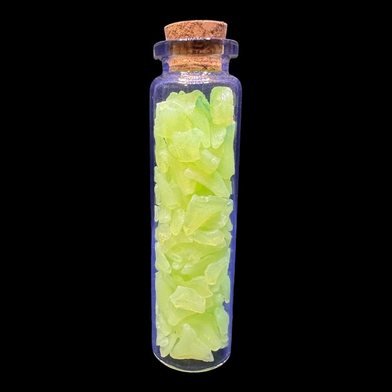 K&ouml;nnte beinhalten: Eine kleine, klare Glasflasche gef&uuml;llt mit hellen, neon-gr&uuml;nen, unregelm&auml;&szlig;ig geformten St&uuml;cken. Die Flasche hat einen Korken und steht vor einem schwarzen Hintergrund. Der Inhalt scheint dekorativ oder f&uuml;r Bastelarbeiten zu sein.