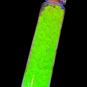 K&ouml;nnte beinhalten: Ein klares Glasfl&auml;schchen gef&uuml;llt mit leuchtend neongr&uuml;nen, unregelm&auml;&szlig;ig geformten Kristallen. Das Fl&auml;schchen hat einen Korken und steht vor einem schwarzen Hintergrund. Die Kristalle scheinen zu leuchten und erzeugen einen auff&auml;lligen visuellen Effekt.