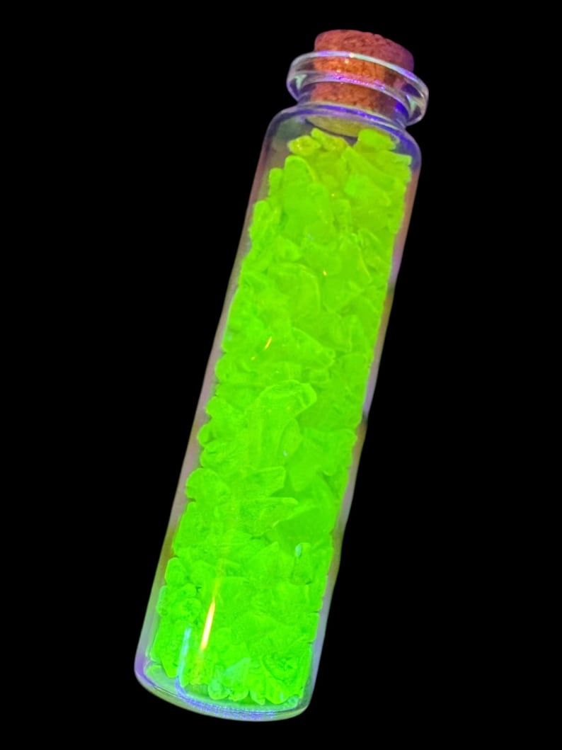 K&ouml;nnte beinhalten: Ein klares Glasfl&auml;schchen gef&uuml;llt mit leuchtend neongr&uuml;nen, unregelm&auml;&szlig;ig geformten Kristallen. Das Fl&auml;schchen hat einen Korken und steht vor einem schwarzen Hintergrund. Die Kristalle scheinen zu leuchten und erzeugen einen auff&auml;lligen visuellen Effekt.