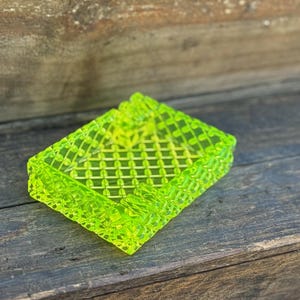 May include: A neon green rectangular ashtray with a lattice design. The ashtray is made of a translucent material and has a textured surface. The ashtray is sitting on a wooden surface.