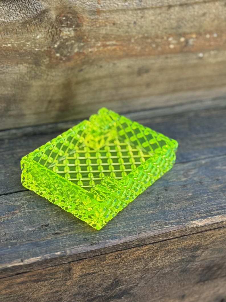 May include: A neon green rectangular ashtray with a lattice design. The ashtray is made of a translucent material and has a textured surface. The ashtray is sitting on a wooden surface.