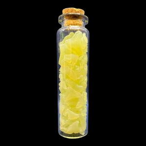 K&ouml;nnte beinhalten: Eine klare Glasflasche mit einem Korken, gef&uuml;llt mit gelb-gr&uuml;nen, unregelm&auml;&szlig;ig geformten St&uuml;cken. Die Flasche ist vor einem schwarzen Hintergrund zentriert. Die St&uuml;cke scheinen eine Art Kristall oder Edelstein zu sein.