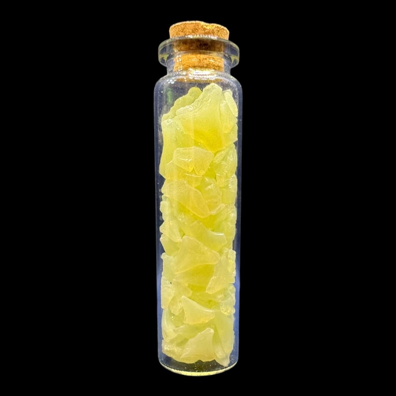 K&ouml;nnte beinhalten: Eine klare Glasflasche mit einem Korken, gef&uuml;llt mit gelb-gr&uuml;nen, unregelm&auml;&szlig;ig geformten St&uuml;cken. Die Flasche ist vor einem schwarzen Hintergrund zentriert. Die St&uuml;cke scheinen eine Art Kristall oder Edelstein zu sein.