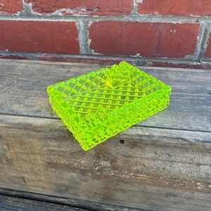 May include: A neon green, rectangular trinket box with a diamond pattern. The box is made of a translucent material and has a lid. The box is sitting on a wooden surface.