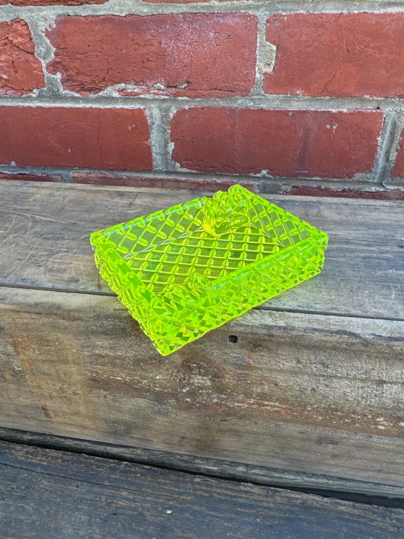 May include: A neon green, rectangular trinket box with a diamond pattern. The box is made of a translucent material and has a lid. The box is sitting on a wooden surface.