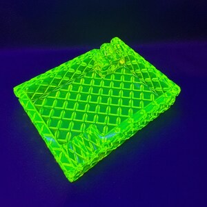 May include: A bright neon green glass trinket box with a diamond pattern. The box has a lid and a decorative handle. The box is sitting on a dark blue surface.