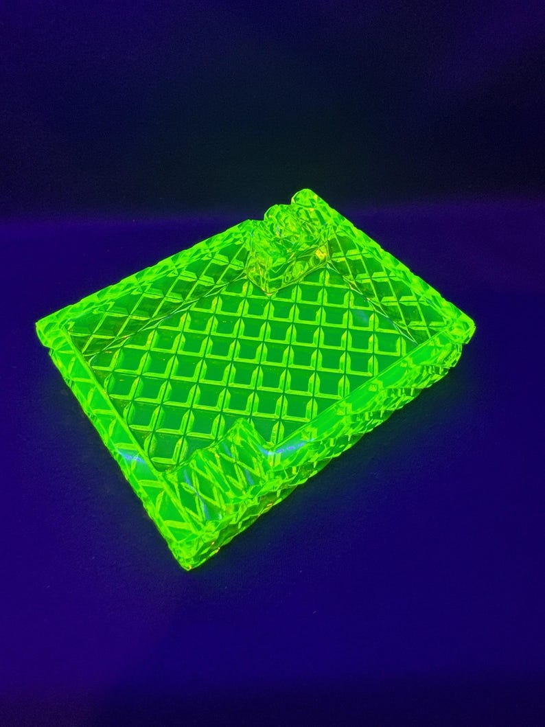 May include: A bright neon green glass trinket box with a diamond pattern. The box has a lid and a decorative handle. The box is sitting on a dark blue surface.
