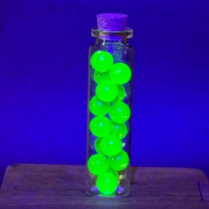 Op de afbeelding: Een doorzichtige glazen fles met een paarse kurkstop gevuld met meerdere kleine, ronde, heldere neongroene bollen die intens oplichten onder ultraviolet licht, wat een levendig contrast vormt met de donkerblauwe achtergrond.