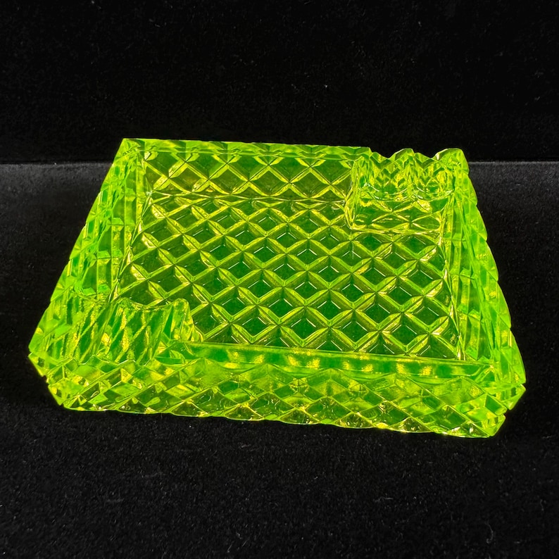May include: A square, neon green ashtray with a diamond pattern. The ashtray has a rectangular shape with a slightly sloped design and a small notch on one side. The glass ashtray is on a black surface.