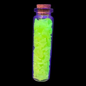 K&ouml;nnte beinhalten: Eine klare Glasflasche gef&uuml;llt mit hellen, neongr&uuml;nen, unregelm&auml;&szlig;ig geformten St&uuml;cken. Die Flasche hat einen Korken und steht vor einem schwarzen Hintergrund. Das Glas hat einen violetten Farbton.