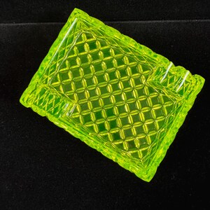 May include: A rectangular, neon green glass ashtray with a diamond pattern. The ashtray has a textured surface and a slightly raised edge. The corners are rounded, and the overall design is geometric.