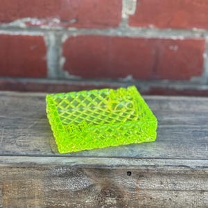 May include: A square, neon green ashtray with a diamond pattern. The ashtray is made of a translucent material and sits on a weathered wooden surface. The background is a red brick wall.