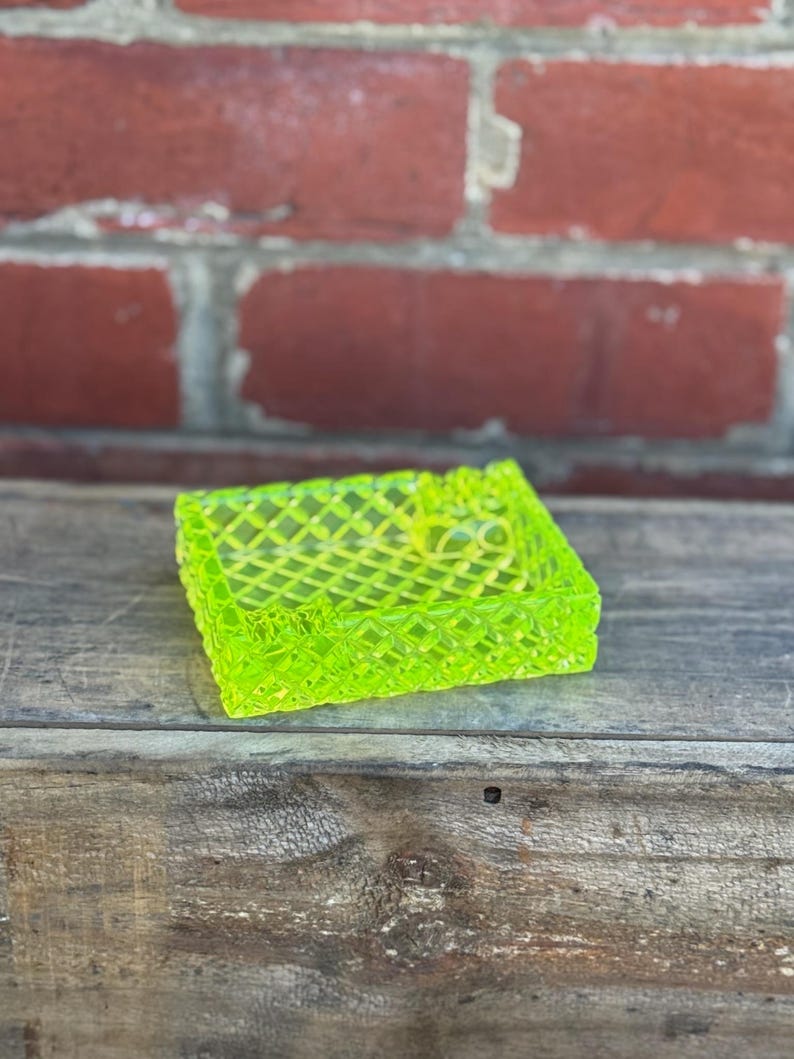 May include: A square, neon green ashtray with a diamond pattern. The ashtray is made of a translucent material and sits on a weathered wooden surface. The background is a red brick wall.