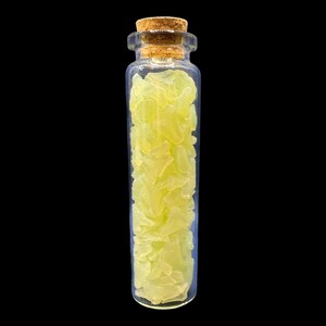 K&ouml;nnte beinhalten: Kleine, klare Glasflasche mit Korken, gef&uuml;llt mit hellgelben, unregelm&auml;&szlig;ig geformten Fragmenten. Die Flasche steht vor einem schwarzen Hintergrund, der den Inhalt hervorhebt. Die Fragmente scheinen kristallin oder mineralisch zu sein.