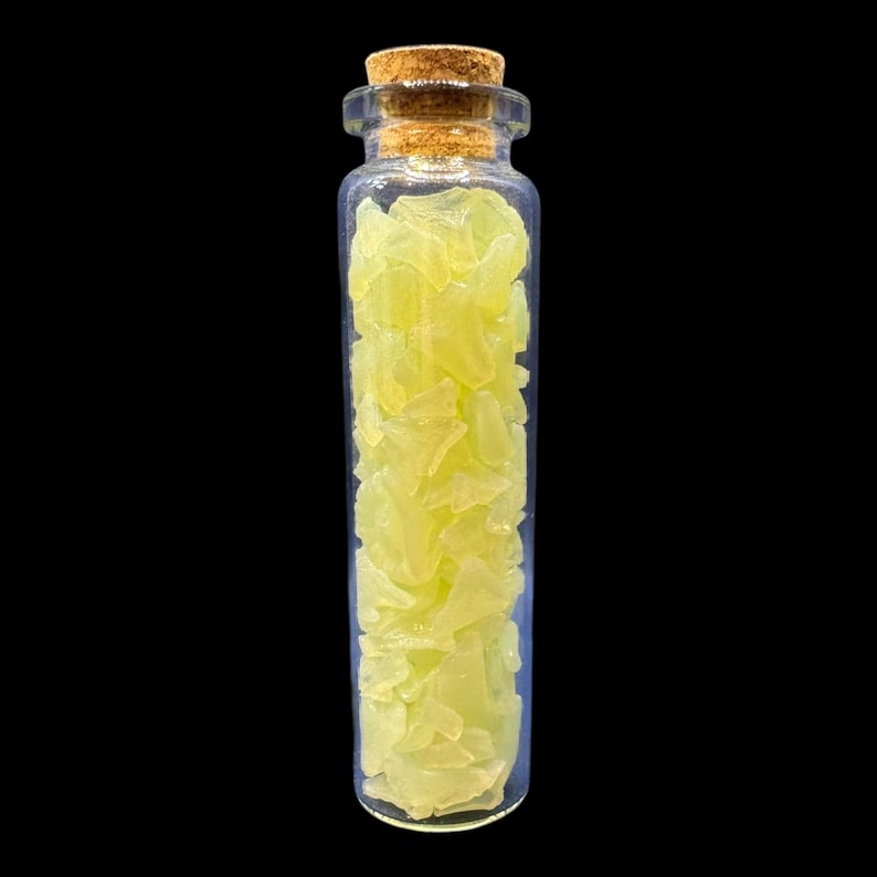 K&ouml;nnte beinhalten: Kleine, klare Glasflasche mit Korken, gef&uuml;llt mit hellgelben, unregelm&auml;&szlig;ig geformten Fragmenten. Die Flasche steht vor einem schwarzen Hintergrund, der den Inhalt hervorhebt. Die Fragmente scheinen kristallin oder mineralisch zu sein.