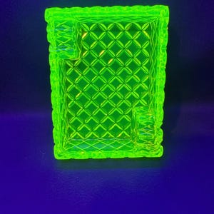 May include: A bright neon green ashtray with a diamond pattern and rectangular shape. The ashtray has a textured rim and two built-in rests for cigarettes. The background is a dark blue.