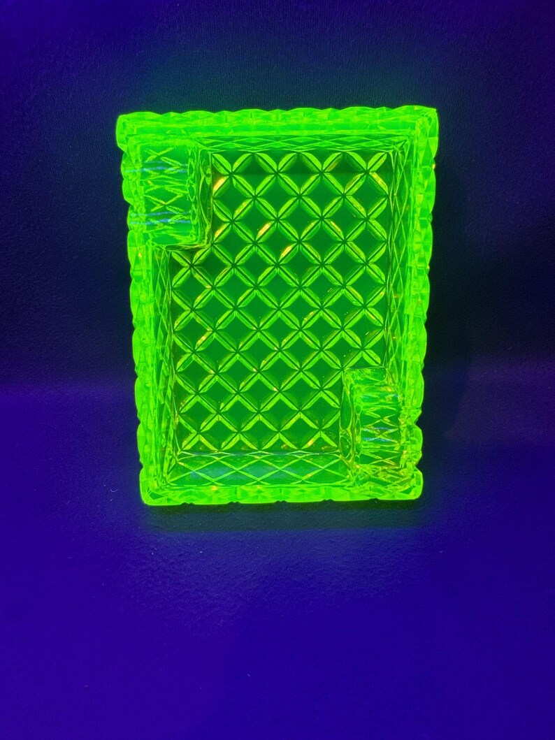 May include: A bright neon green ashtray with a diamond pattern and rectangular shape. The ashtray has a textured rim and two built-in rests for cigarettes. The background is a dark blue.