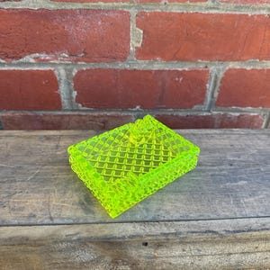May include: A neon green, rectangular trinket box with a diamond pattern. The box has a lid and is made of a translucent material. The box is sitting on a wooden surface.