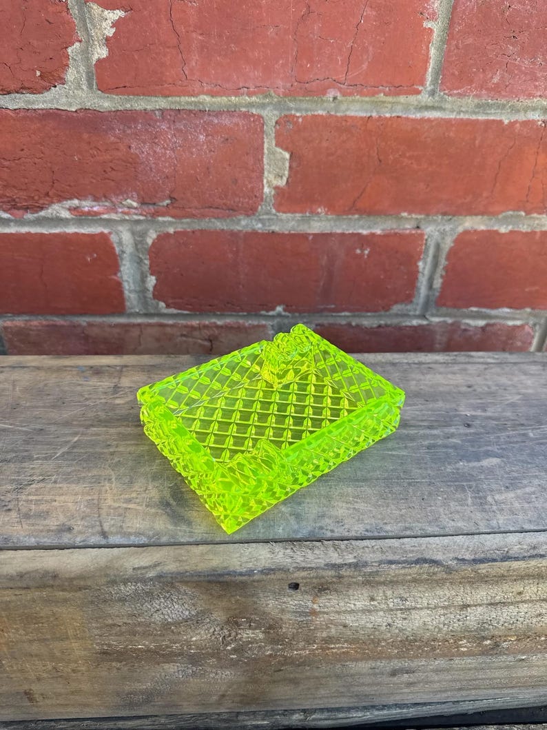 May include: A neon green, rectangular trinket box with a diamond pattern. The box has a lid and is made of a translucent material. The box is sitting on a wooden surface.