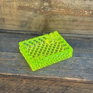 May include: A neon green, rectangular trinket box with a diamond pattern and a small floral accent on the lid. The box is made of a translucent material and sits on a dark wooden surface.