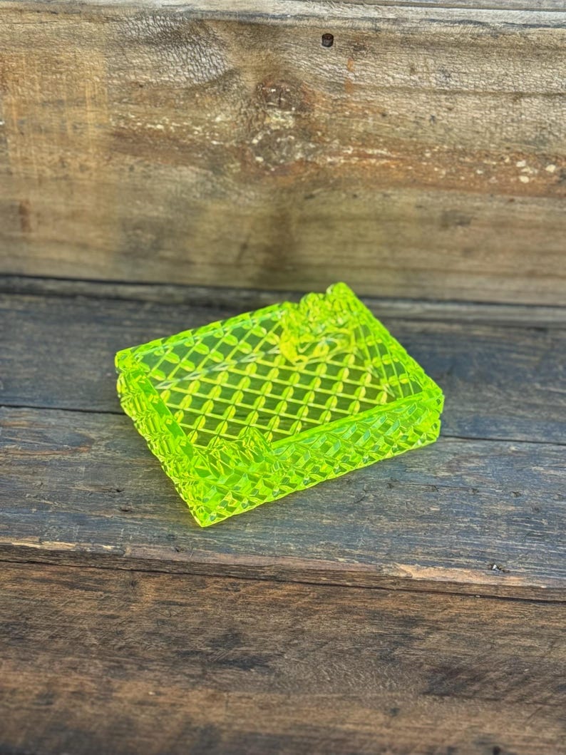 May include: A neon green, rectangular trinket box with a diamond pattern and a small floral accent on the lid. The box is made of a translucent material and sits on a dark wooden surface.