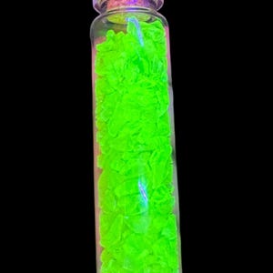 K&ouml;nnte beinhalten: Eine kleine, klare Glasflasche gef&uuml;llt mit leuchtend neongr&uuml;nen Fragmenten. Die Flasche ist mit einem Korken verschlossen. Der Hintergrund ist schwarz und betont den leuchtenden gr&uuml;nen Inhalt. Der Inhalt der Flasche scheint zu leuchten.