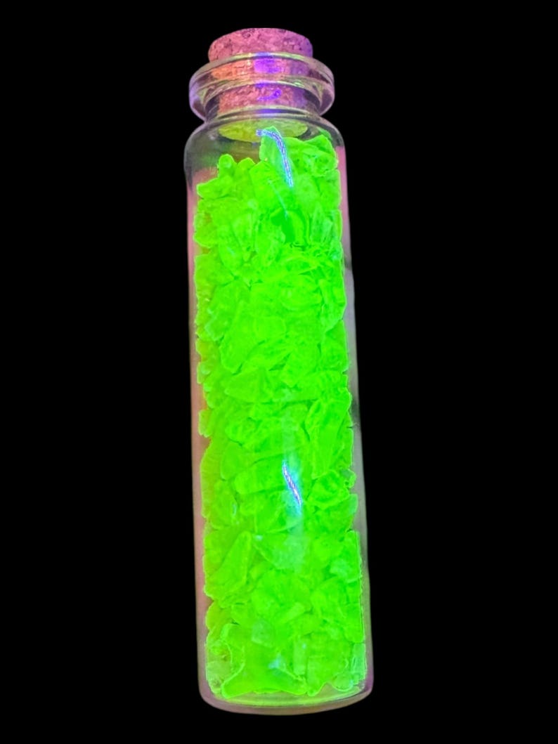 K&ouml;nnte beinhalten: Eine kleine, klare Glasflasche gef&uuml;llt mit leuchtend neongr&uuml;nen Fragmenten. Die Flasche ist mit einem Korken verschlossen. Der Hintergrund ist schwarz und betont den leuchtenden gr&uuml;nen Inhalt. Der Inhalt der Flasche scheint zu leuchten.