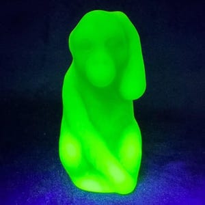 May include: A glowing green figurine of a seated figure with arms crossed. The sculpture has a smooth, rounded form and is illuminated against a dark blue background. The figurine appears to be made of glass or a similar material.