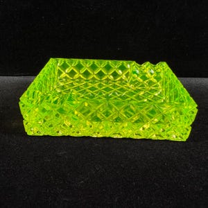 May include: A neon green ashtray with a geometric diamond pattern. The rectangular ashtray has a textured surface and a small notch on one side. The ashtray is made of glass and has a bright, eye-catching color.