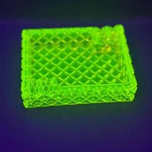 May include: A square, neon green ashtray with a diamond pattern. The ashtray has a raised edge and a small rest for a cigarette. The object is made of glass and sits on a dark blue surface.