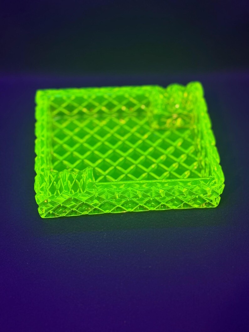 May include: A square, neon green ashtray with a diamond pattern. The ashtray has a raised edge and a small rest for a cigarette. The object is made of glass and sits on a dark blue surface.
