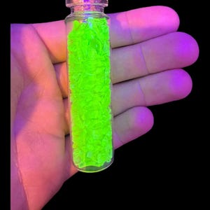 K&ouml;nnte beinhalten: Eine kleine, klare Glasflasche gef&uuml;llt mit hellen, neon-gr&uuml;nen, unregelm&auml;&szlig;ig geformten St&uuml;cken. Die Flasche hat einen Korken. Der Hintergrund ist schwarz, und die Flasche wird in einer Hand gehalten, wobei die Haut durch rosa und violettes Licht beleuchtet wird.