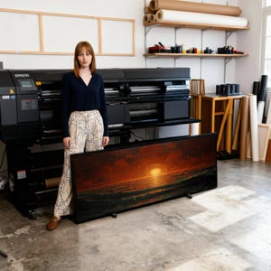 Könnte beinhalten: Ein großes, rechteckiges, gerahmtes Kunstwerk, das einen Sonnenuntergang über dem Meer zeigt. Das Kunstwerk weist Farbtöne von Orange, Rot und Dunkelgrün auf. Der Rahmen ist schwarz. Das Kunstwerk wird in einem Studio mit einem großen Drucker und Kunstbedarf ausgestellt.