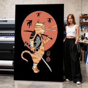 Könnte beinhalten: Ein großer schwarzer Leinwanddruck mit einem Cartoon-Katzen-Samurai mit Schwertern, einem Fischtattoo und einem roten Sonnenhintergrund. Die Katze trägt ein Stirnband und Bandagen. Der Druck wird in einer Werkstatt gezeigt.