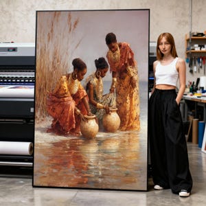 Op de afbeelding: Grote ingelijste kunstdruk met drie vrouwen in traditionele kledij bij water. Ze interageren met grote kleipotten. Het kunstwerk gebruikt warme tinten oranje, goud en bruin, met een reflecterend wateroppervlak.