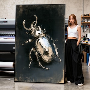 May include: A large, framed artwork featuring a metallic silver beetle against a dark, textured background. The beetle is highly detailed, with reflective surfaces. The artwork is displayed in a studio setting.