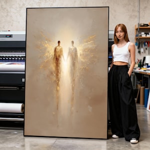 Könnte beinhalten: Großes abstraktes Gemälde in einem schwarzen Rahmen. Das Kunstwerk zeigt zwei Figuren, die sich an den Händen halten, mit goldenen Flügeln und hellem Licht dahinter. Die Farbpalette ist hauptsächlich Beige und Gold.