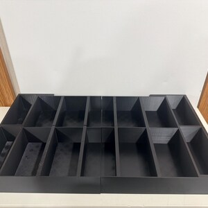 May include: A black, multi-compartment organizer with rectangular and angled sections. The organizer is made of a dark material and is designed to hold various items. The compartments are arranged in rows and columns.