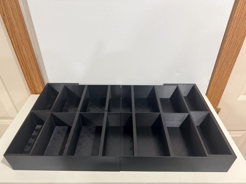 May include: A black, multi-compartment organizer with rectangular and angled sections. The organizer is made of a dark material and is designed to hold various items. The compartments are arranged in rows and columns.