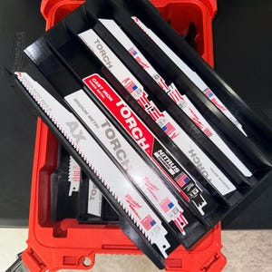 May include: A red toolbox with black inserts holding several reciprocating saw blades. The blades are white with black and red text, including "TORCH" and "SAWZALL". Some blades have the American flag printed on them.